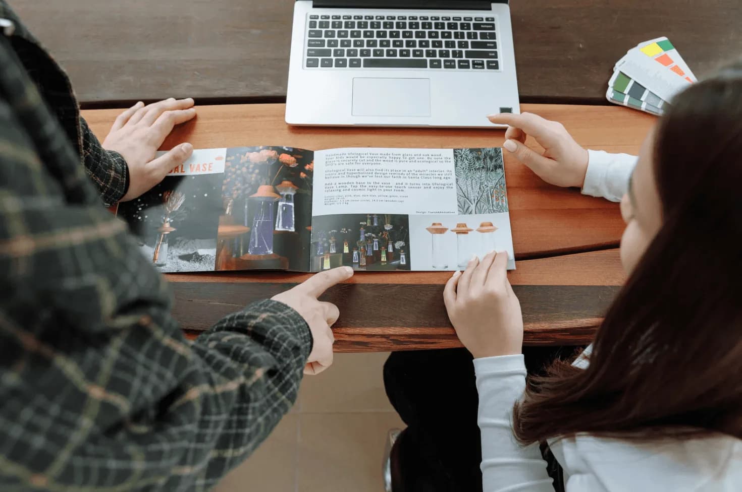 Two people reviewing a printed product catalogue with a laptop and swatch palette on the table.