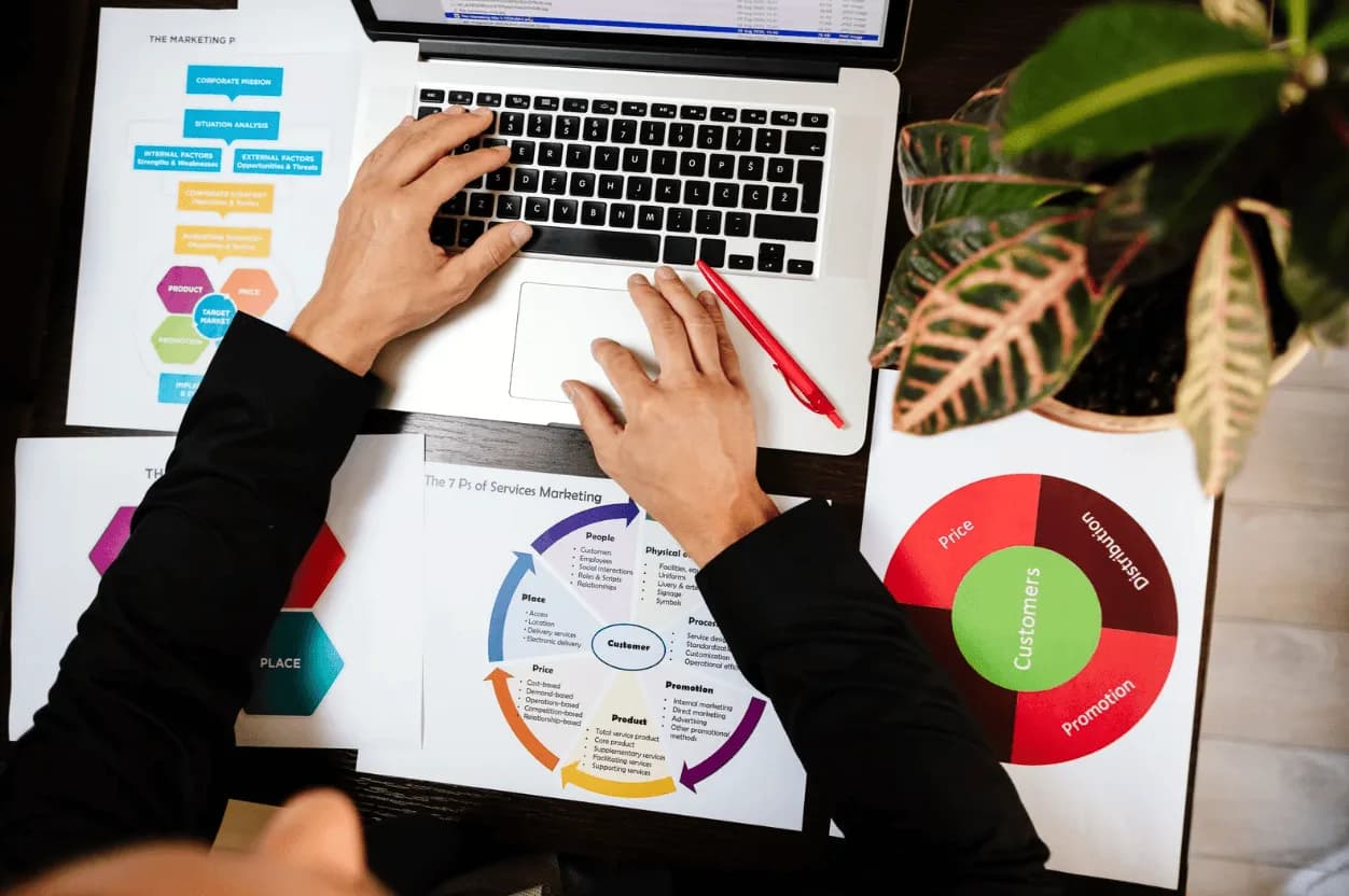 Marketer analysing strategy documents on laptop surrounded by marketing diagrams — highlighting keyword planning and campaign execution.