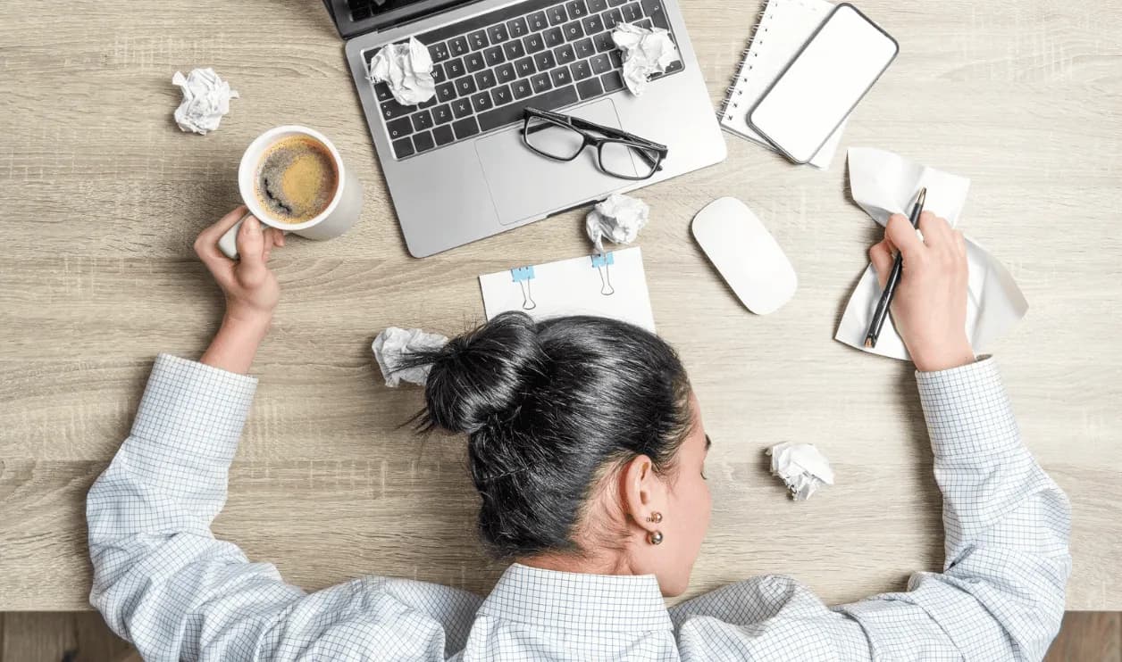 Exhausted marketer with head on desk surrounded by coffee and crumpled papers — showing the chaos of manual campaign scaling without automation.
