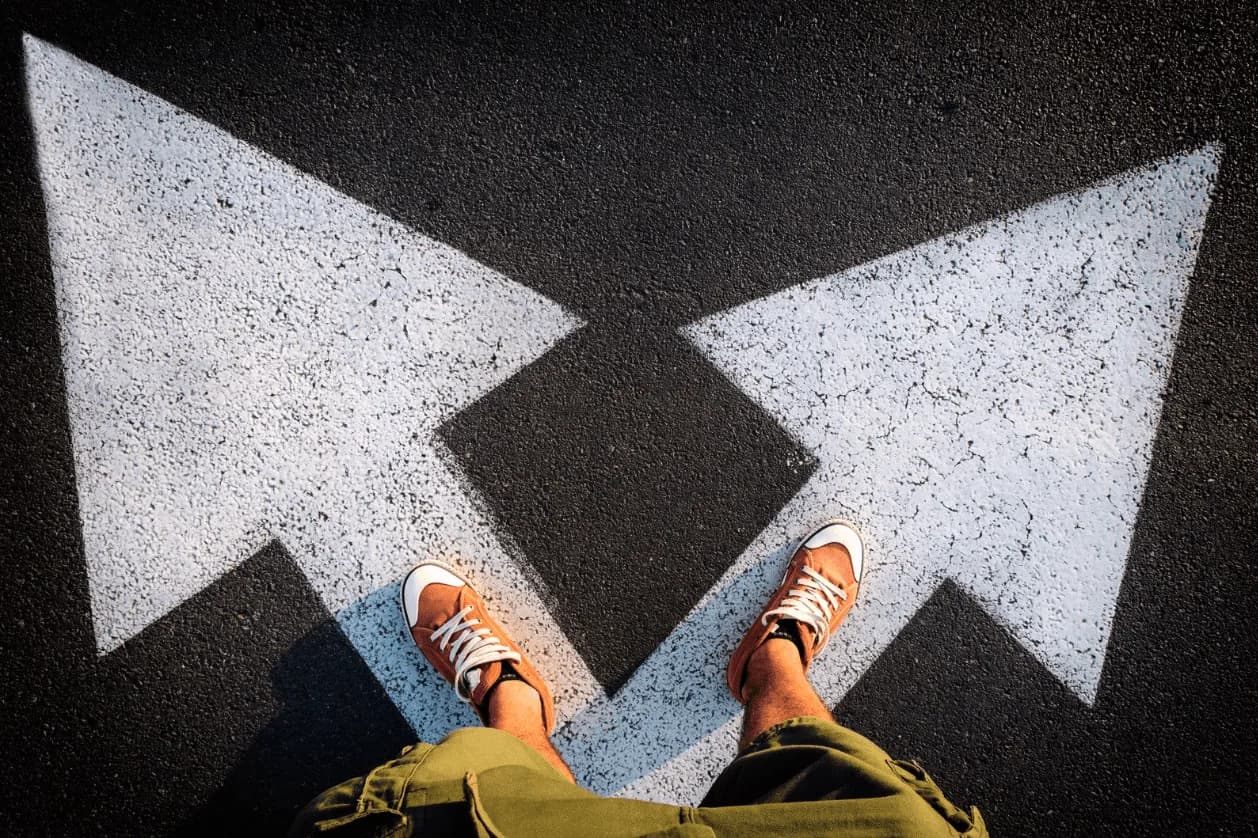 Person standing at a crossroad with two large white arrows painted on asphalt, symbolising strategic decision-making in marketing direction.