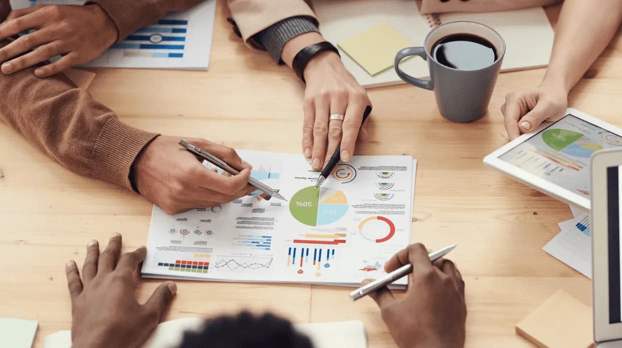 Team members pointing at printed sales charts and a tablet with performance graphs, representing collaborative Amazon portfolio analysis to improve profitability.