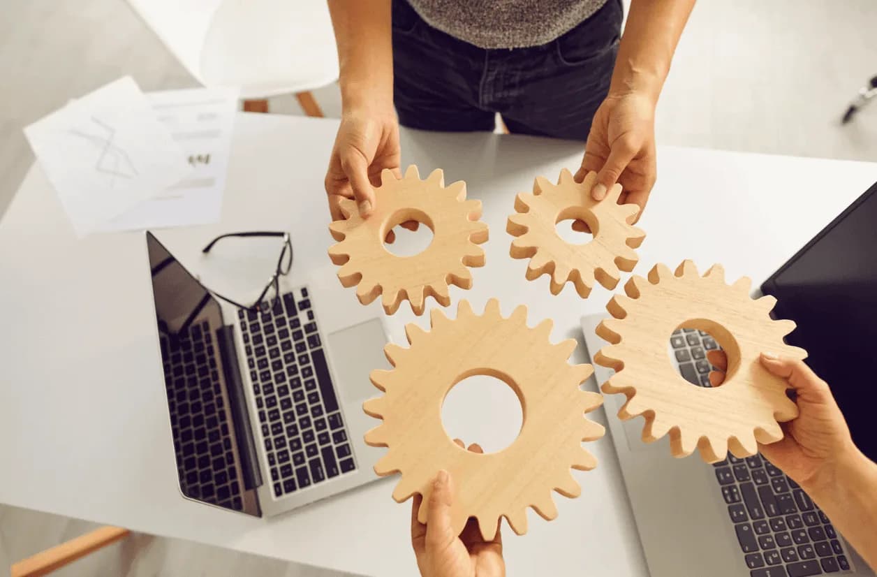 Three people holding interlocking wooden gears over a table with laptops and documents, symbolising teamwork and collaboration.